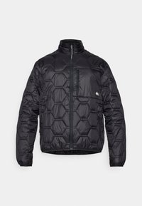 Sort pufferjakke med en hexagonal quiltet design, høj krave, lynlås foran og en brystlomme med lynlås.