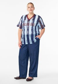 Blau-weiß gestreifter Spitzentop mit kastanienbraunen Akzenten und dem Text "MAGNIFIQUE", kombiniert mit lockeren dunklen Jeanshosen und dunklen flachen Schuhen.
