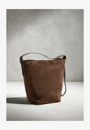 Ruda zomšinė kibirinė rankinė su vienu petnešos dirželiu, padėta ant neutralios pilkos ir baltos spalvų fono.