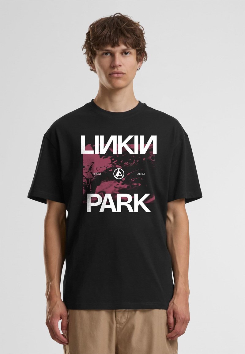 Jeune homme aux cheveux bouclés portant un t-shirt noir du groupe Linkin Park et un pantalon beige, debout devant un fond clair uni.