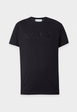 Jonge man staat met handen in de zakken, draagt een zwart reliëf T-shirt, lichtblauwe spijkerbroek en zwarte nette schoenen op een effen witte achtergrond.