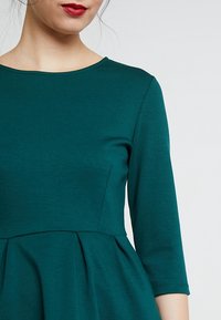 Teal jurk met driekwart mouwen, ronde halslijn en een aansluitende pasvorm die uitloopt in een geplooide rok. Soepele textuur, minimalistisch ontwerp.
