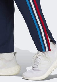 Marineblauwe sweatpants met rood, wit en blauwe zijkanten, voorzien van een zijspleet. Gecombineerd met witte sportschoenen.