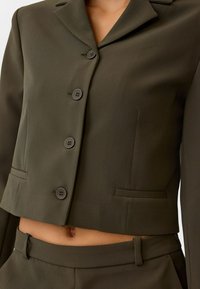 Blazer court vert olive avec un col cranté, fermeture à quatre boutons et deux poches avant. Texture lisse, coupe ajustée.