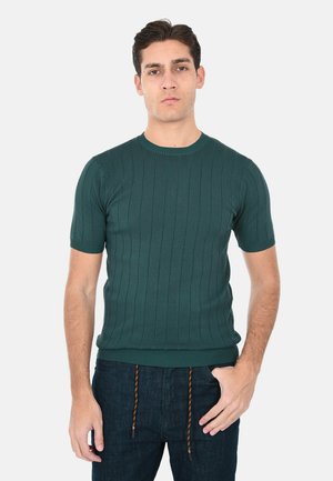 Uomo in piedi, volto frontale, che indossa un maglione a maniche corte a coste di colore verde scuro e pantaloni scuri con coulisse marroni, su uno sfondo chiaro e uniforme.
