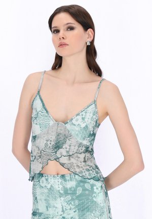 Donna che indossa un top corto senza maniche con motivi e spalline sottili, abbinato a una gonna, in piedi con le mani dietro la schiena su uno sfondo bianco.