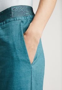 Close-up van een hand die rust in de zak van een blauwe broek met een gemêleerde donkergroene tailleband.