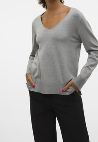 Šedý svetr s výstřihem do V z měkké tkaniny, s dlouhými rukávy a žebrovanými manžetami. Model má na sobě černé kalhoty, ruce v kapsách.