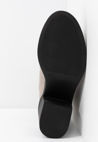 Semelle d'une botte beige à talon et cheville avec semelle extérieure en caoutchouc noir texturé et talon bloc sur fond blanc.