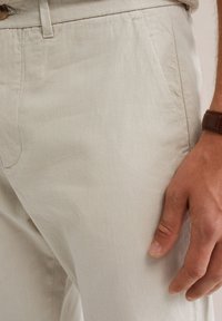 Pantaloni beige chiaro a fantasia con una sottile trama a quadri, dotati di tasche laterali e chiusura con bottone, mostrati con una mano appoggiata nei pressi.