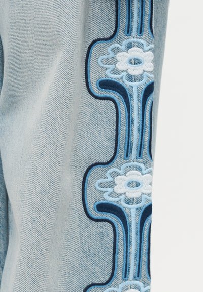 Ανοιχτό μπλε τζιν ύφασμα με κάθετη λουλουδάτη κεντητική σε άσπρο και αποχρώσεις του μπλε κατά μήκος της ραφής.