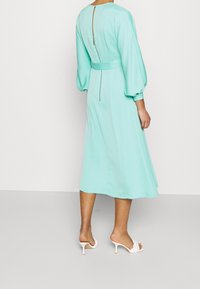 En mintgrön, knälång klänning med långa puffärmar, en figurnära midja och en dragkedjedetalj i ryggen. Kombinerad med vita klacksandaler.