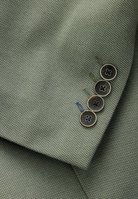 Zielony, teksturowany materiał z subtelnym wzorem, z czterema czarnymi guzikami w kwiatowe wzory oraz kontrastującymi detalami wyszycia.