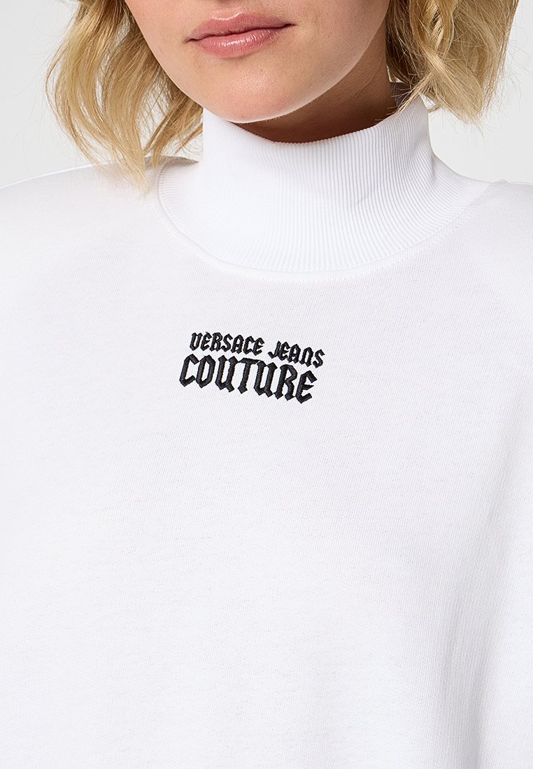 Kobieta ubrana w biały sweter z golfem w prążki, z haftem "Versace Jeans Couture" w kolorze czarnym na klatce piersiowej.