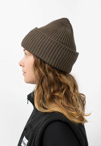 Bruine gebreide beanie met een geribbelde textuur en een puntige top, gedragen door een persoon met golvend, schouderlang haar. Zwarte vest eronder.