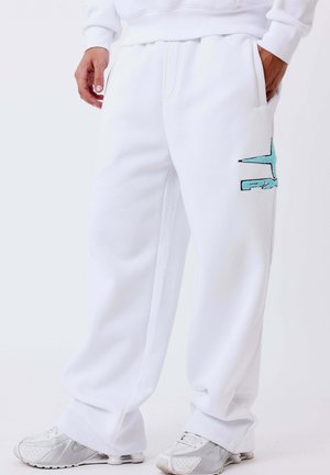 Personne portant un pantalon de survêtement blanc avec un logo bleu sur la jambe et des baskets blanches, debout avec une main dans la poche.