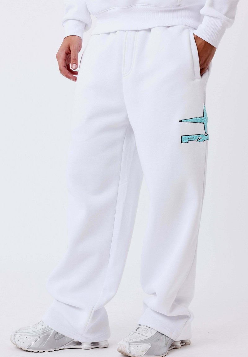 Personne portant un pantalon de survêtement blanc avec un logo bleu sur la jambe et des baskets blanches, debout avec une main dans la poche.