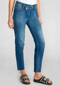 Skinny blue jeans made of denim, featuring a mid-rise design, contrasting stitching, and a classic five-pocket style. Paired with black sandals.