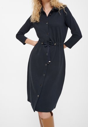 Vrouw die een donkerblauw overhemdjurk met knopen draagt, met lange mouwen, een taillecord en beige kniehoge laarzen.