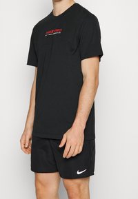 Musta Nike Pro t-paita lyhyillä hihoilla ja punaisella logolla rinnassa, yhdistettynä mustiin shortseihin, joissa on valkoinen Nike swoosh. Sileä kangastuntuma.