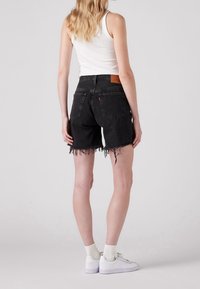 Pantalones cortos de mezclilla negra con dobladillo deshilachado, bolsillos traseros y una etiqueta de cuero marrón claro. Combinados con una camiseta sin mangas blanca y zapatillas blancas.