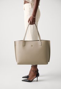 Beige leren tote handbag met twee platte handvatten, een gladde textuur en een gegraveerd logo. Gecombineerd met aangesloten lichtgekleurde broek en zwarte hakken.