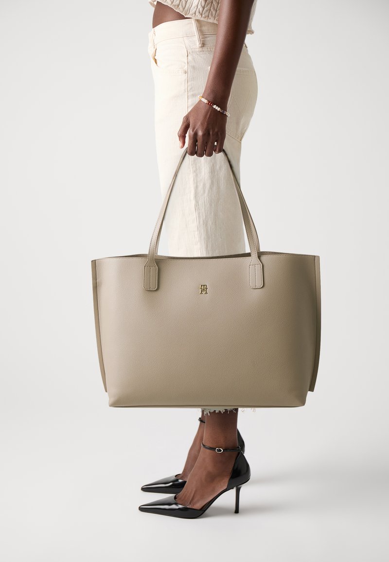 Beige leren tote handbag met twee platte handvatten, een gladde textuur en een gegraveerd logo. Gecombineerd met aangesloten lichtgekleurde broek en zwarte hakken.