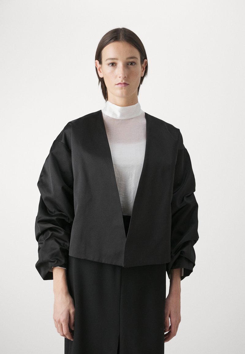 Black cropped jacket with oversized sleeves, made of smooth fabric. Layered over a sheer white top, featuring a high neck and slim design.