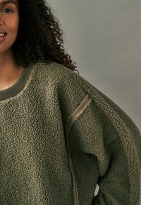 Oliven grøn fleece sweatshirt med en tekstureret overflade, brede ærmer og en ribbet halsudskæring med synlige sømdetaljer.
