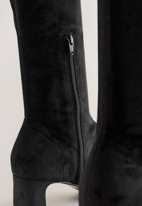 Bottes hautes jusqu'au genou en daim noir, dotées d'une fermeture éclair latérale et d'un talon haut effilé. Texture lisse avec des détails de couture subtils.