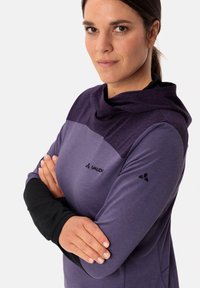 Maglia lunga viola con collo a drappeggio, caratterizzata da una sezione superiore più scura, maniche nere e un logo sul petto e sul braccio.