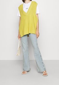 Limetkově zelená pletená vesta s hlubokým V výstřihem, nošená přes bílé tričko. V kombinaci se světle modrými rozšířenými džíny a bílými sandály na pásky.