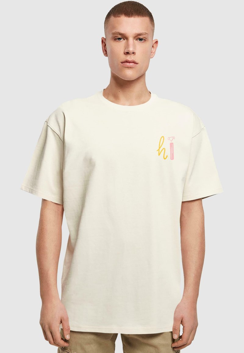 Beżowa bawełniana koszulka, luźny krój, krótkie rękawy. Na lewej stronie klatki piersiowej znajduje się kolorowy napis "hi" w żółtym i różowym kolorze.