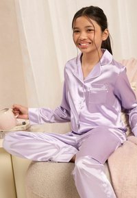 Conjunto de pijama de satén lavanda con botones en la parte delantera, detalles de bolsillo y acentos de ribete blanco, acompañado de una taza rosa sobre un sofá acogedor.