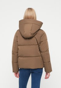 Casaco puffer castanho com capuz, apresentando um design acolchoado e silhueta elongada, usado sobre jeans, visto de costas.