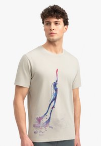 Beige katoenen t-shirt met een kleurrijk abstract ontwerp van een figuur in blauw en roze, voorzien van een ronde halslijn en korte mouwen.