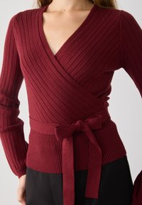 Sweater envolvente bordeaux com riscas, com decote em V, mangas ajustadas e um cinto amarrado na cintura. Tecido texturizado e padrões de riscas contrastantes.