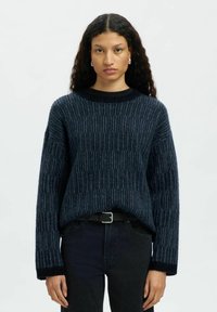 Sieviete ar gariem viļņotiem matiem, ģērbusies zilā tekstūrētā adījumā, ar melnu jostu un tumšiem džinsiem, stāv uz vienkārša gaiša fona.