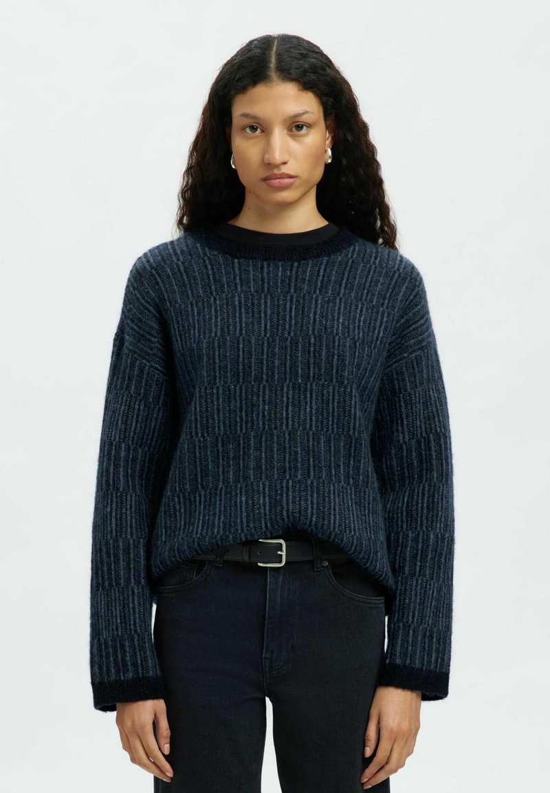 Sieviete ar gariem viļņotiem matiem, ģērbusies zilā tekstūrētā adījumā, ar melnu jostu un tumšiem džinsiem, stāv uz vienkārša gaiša fona.