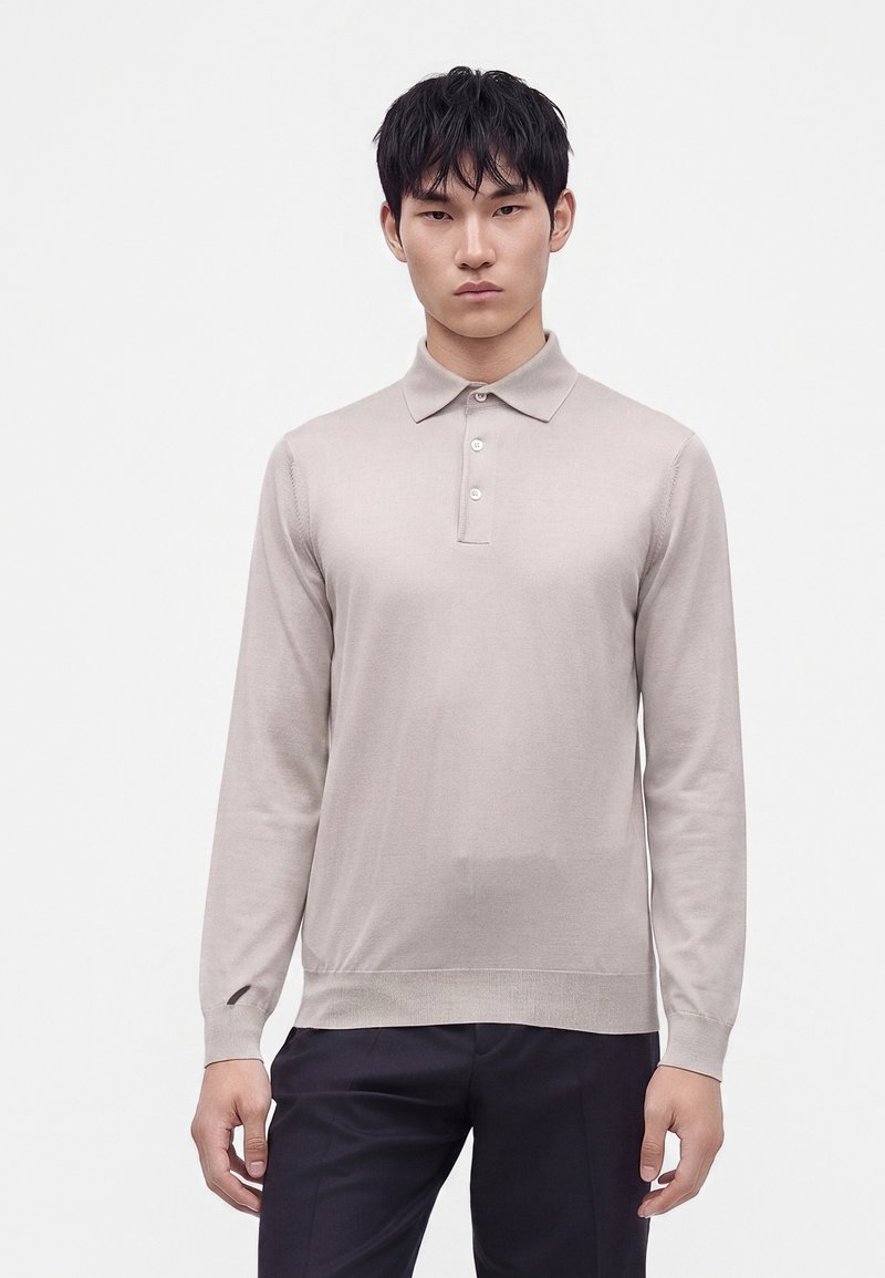 Jeune homme aux cheveux courts et noirs, portant un polo beige clair à manches longues et un pantalon foncé, debout devant un fond blanc uni.