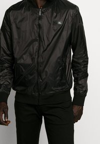 Czarna bomberka wykonana z błyszczącego materiału, z zamkiem z przodu, ściągaczami na mankietach i w pasie, z naszywką z logo na piersi.