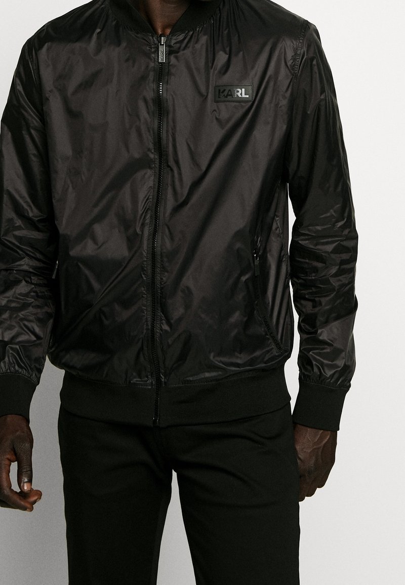 Czarna bomberka wykonana z błyszczącego materiału, z zamkiem z przodu, ściągaczami na mankietach i w pasie, z naszywką z logo na piersi.