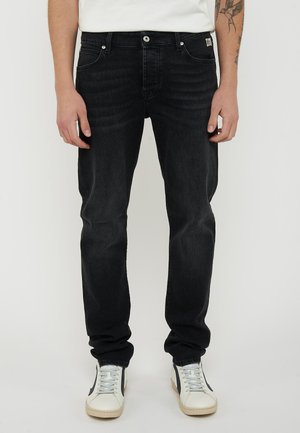 Uomo che indossa jeans neri e sneakers bianche con design nero, in piedi davanti a uno sfondo chiaro e uniforme.