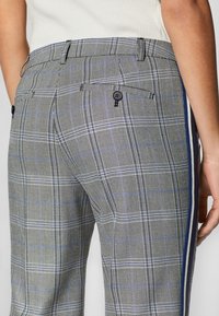 Grijze geruite broek met een prominent ruitpatroon met blauwe accenten, dubbele achterzakken en een gladde textuur. Zijstrepen voegen detail toe.