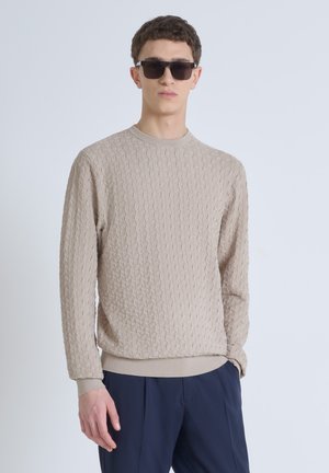 Jersey de punto texturizado beige con cuello redondo y puños acanalados, combinado con pantalones oscuros y gafas de sol negras. Fondo liso.