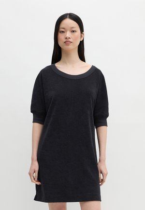 Kobieta stojąca przodem, ubrana w czarną sukienkę do kolan z rękawami do łokcia i szerokim dekoltem.