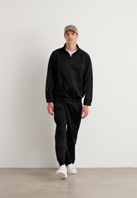 Fato de treino preto com casaco com fecho zip e calças com cintura elástica, feito de tecido suave. Modelo a usar um boné cinza claro e sapatilhas brancas.