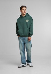 Green hooded sweatshirt with a kangaroo pocket and front graphic; paired with light blue jeans and black and white sneakers.