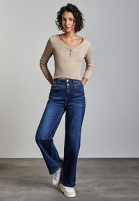 Haut henley beige à manches longues et jean taille haute bleu foncé, associé à des baskets blanches, porté par un modèle sur fond uni.