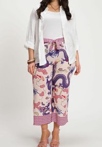 Wijde broek in pastelroze met een draak- en bloemenpatroon in blauw en rood, met een patroon op de tailleband en zoom. Gecombineerd met een wit overhemd.